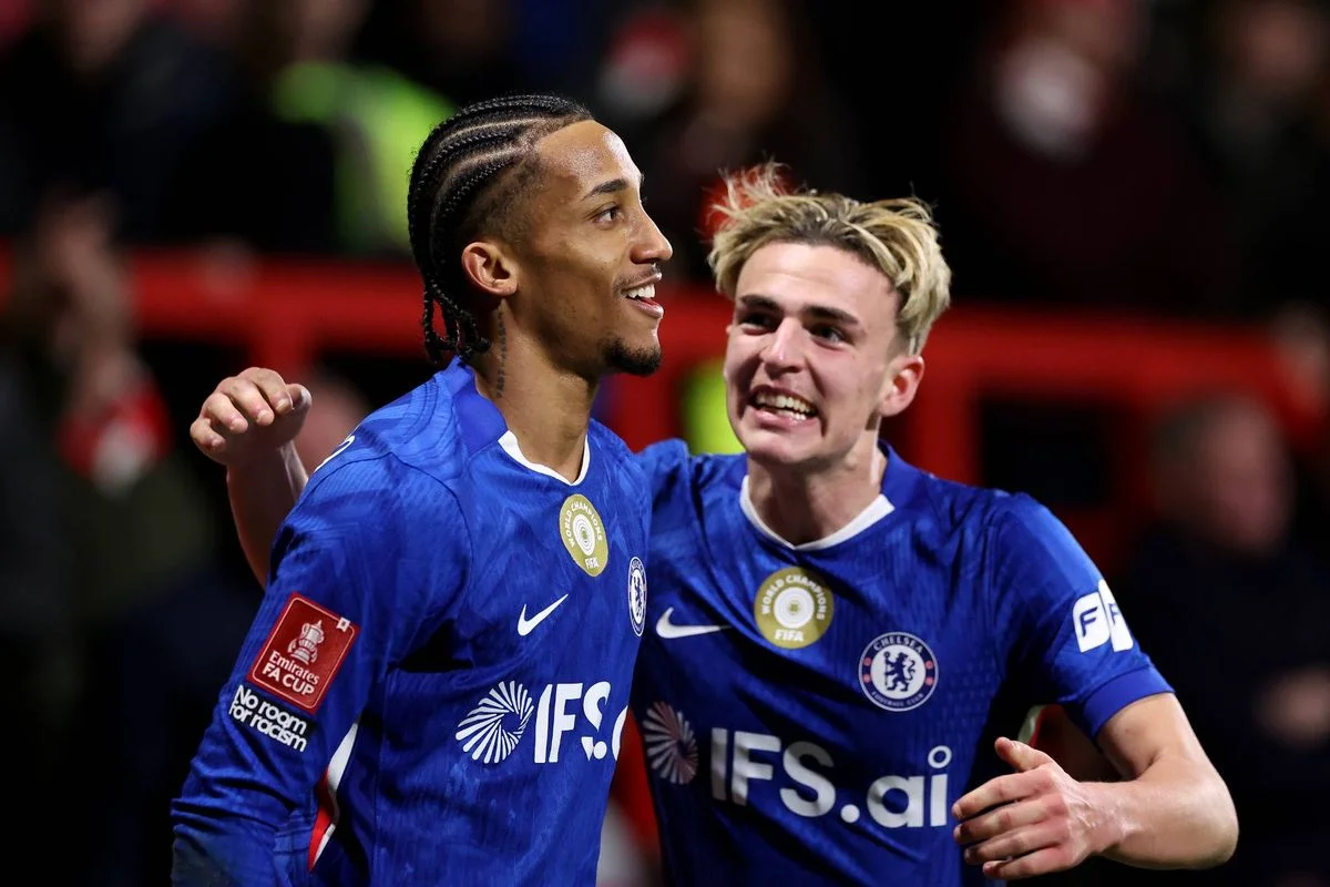 Two male soccer players in blue uniforms celebrate on the field, standing close to each other with their arms around each other, in front of a blurry background of spectators.